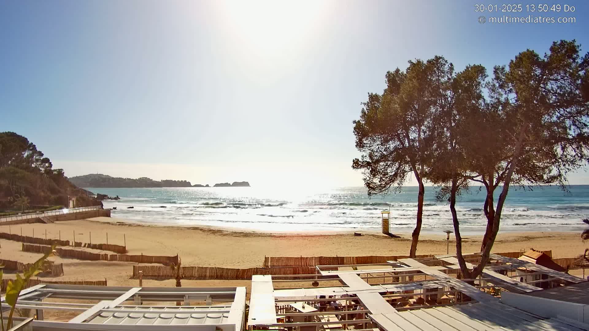 A sandy beach with gentle waves under a sunny sky is viewed from above a partially shaded outdoor patio area, with trees and a lifeguard stand visible in the distance.