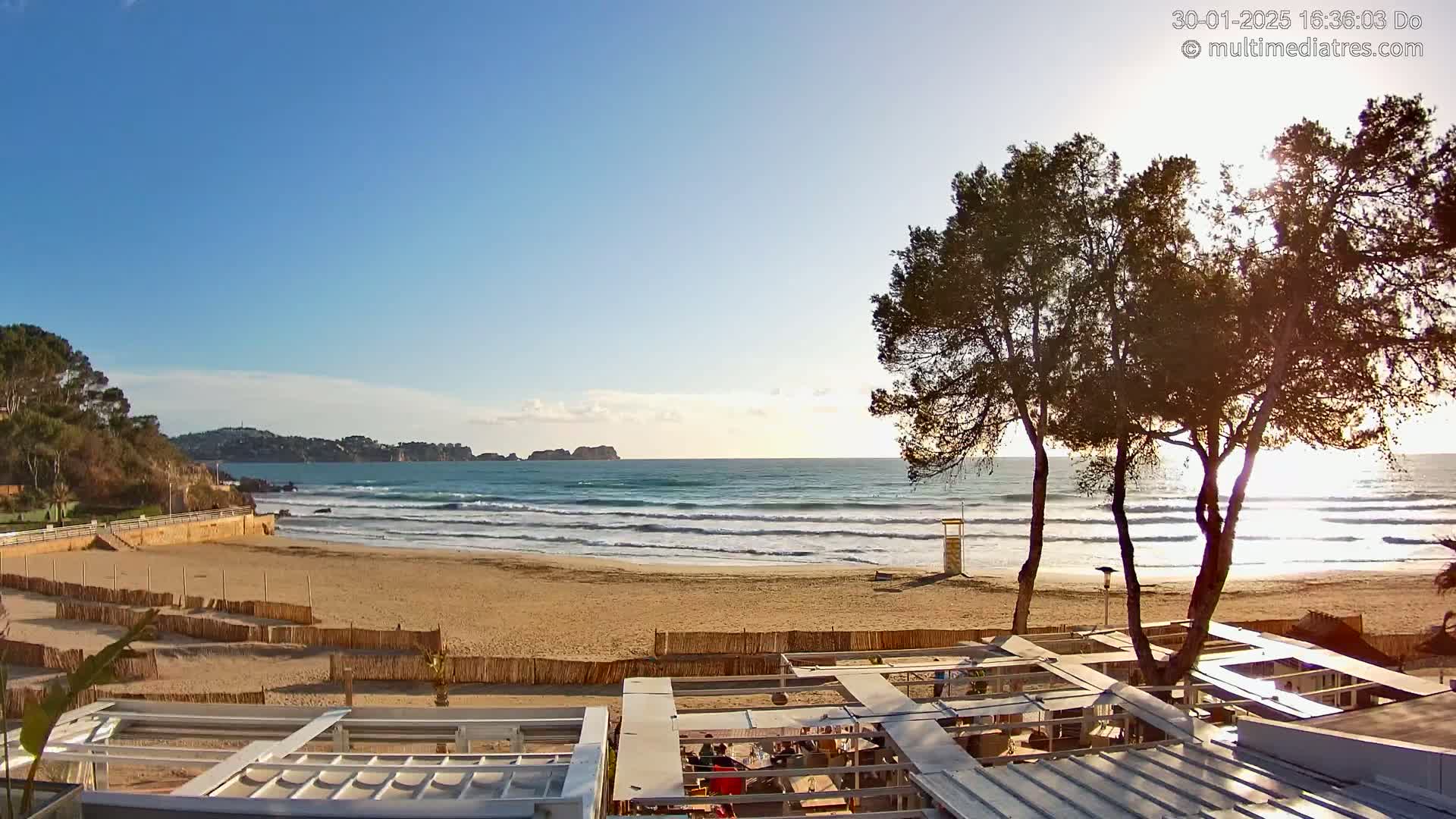 A sandy beach with gentle waves under a clear blue sky is partially visible from behind a beachfront restaurant; several trees stand between the viewer and the ocean.