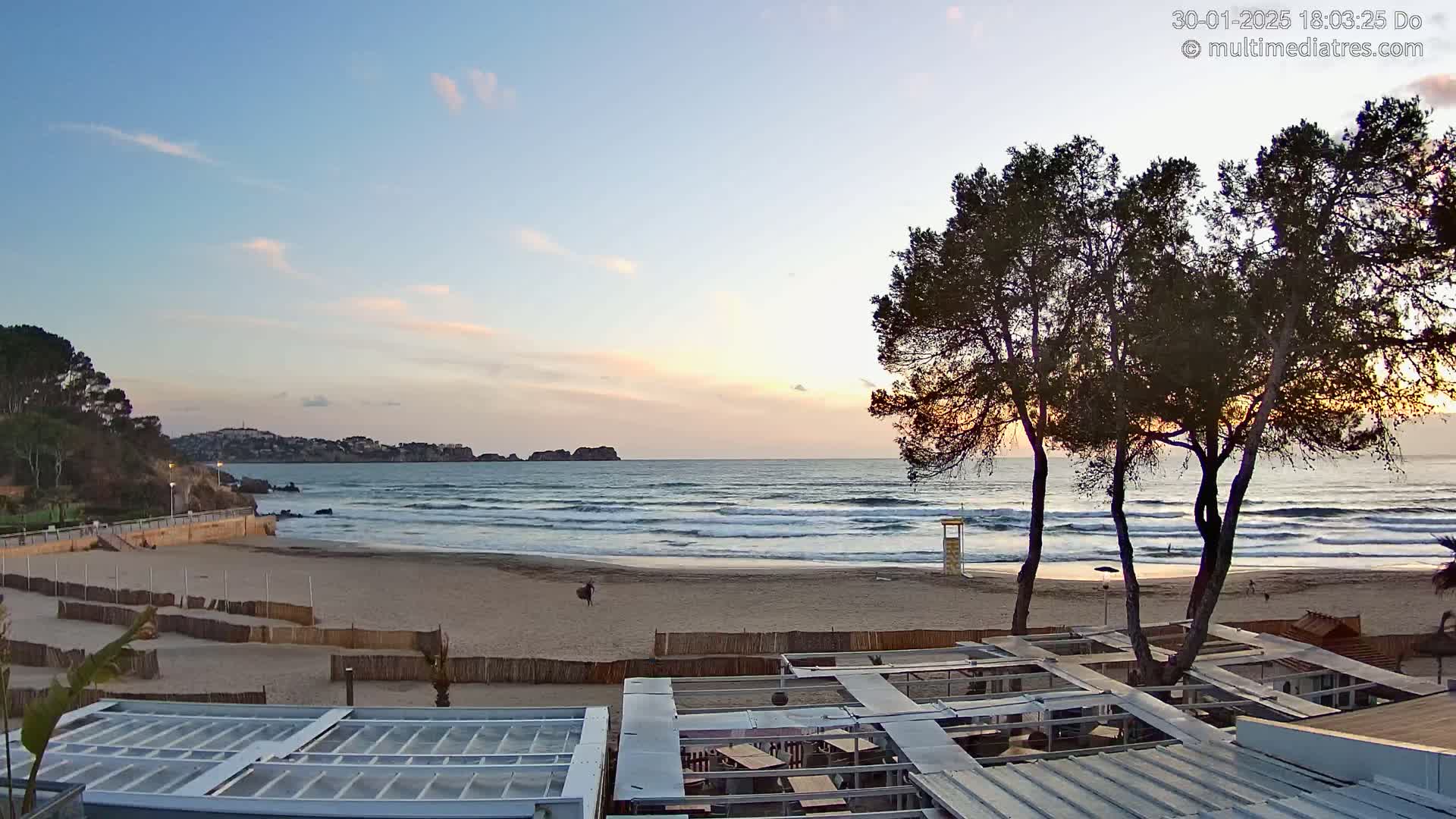 A sandy beach with gentle waves under a mostly clear sunset sky, bordered by trees and beachfront structures.