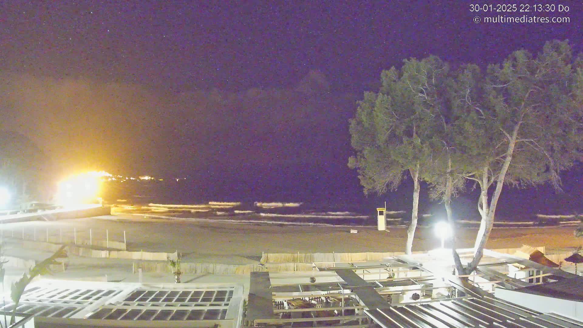 A nighttime view of a beach with calm waves under a starry sky, showing a sandy shore, a beachfront structure with outdoor seating, and two tall trees.