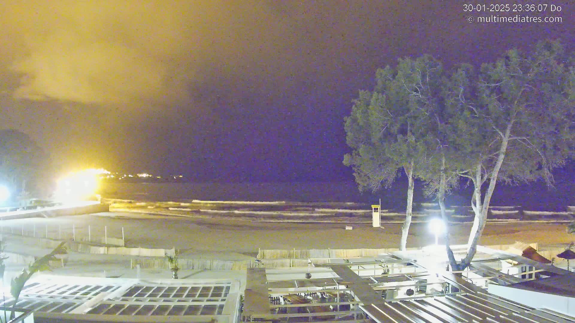 A nighttime beach scene shows a sandy shore with gentle waves under a hazy, yellowish sky, overlooked by a patio and several trees.