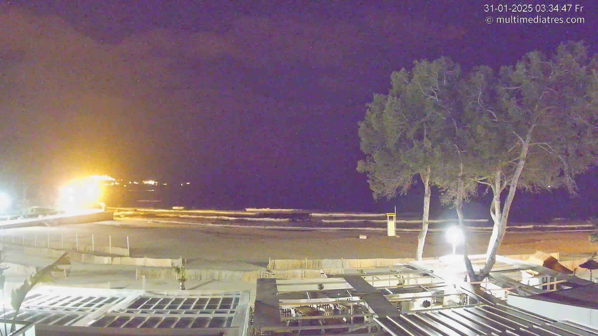 A nighttime beach scene shows a dark sandy beach with gentle waves under a clear, dark sky, and a partially visible building with outdoor seating in the foreground, illuminated by artificial lighting.