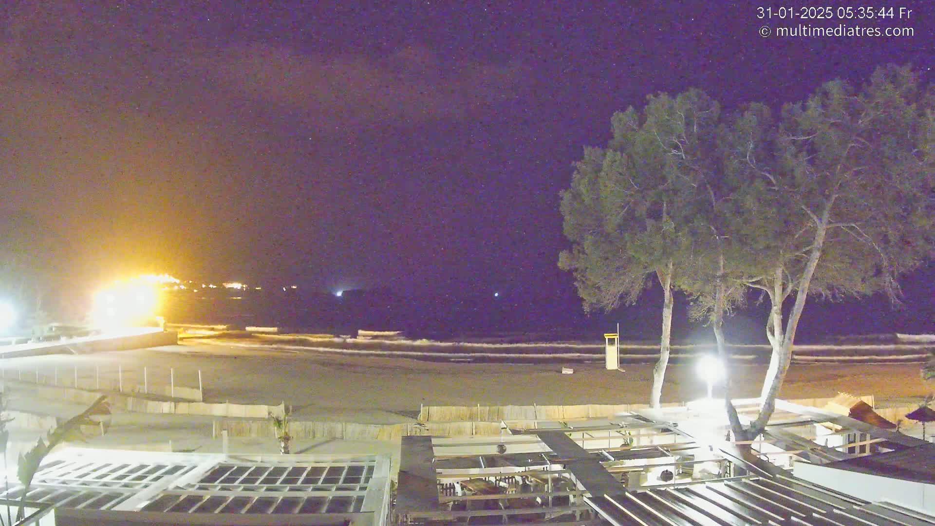 A nighttime view of a beach with gentle waves, illuminated by lights from a beachfront structure under a starry sky.
