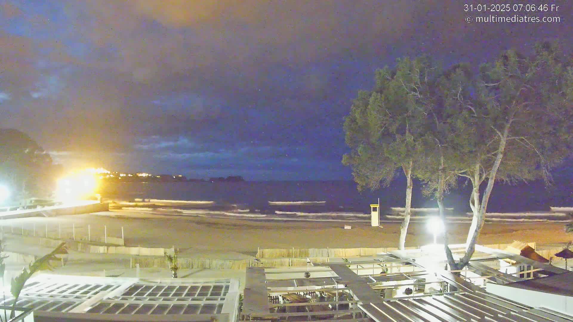 A nighttime view of a sandy beach with gentle waves, under a mostly clear, dark blue sky, shows an outdoor restaurant patio area with trees in the foreground.