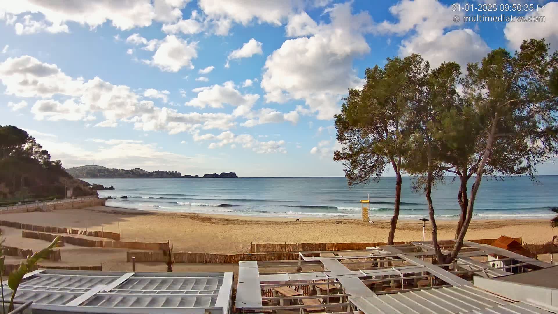 A sandy beach with gentle waves under a partly cloudy sky is viewed from a seaside establishment with outdoor seating.