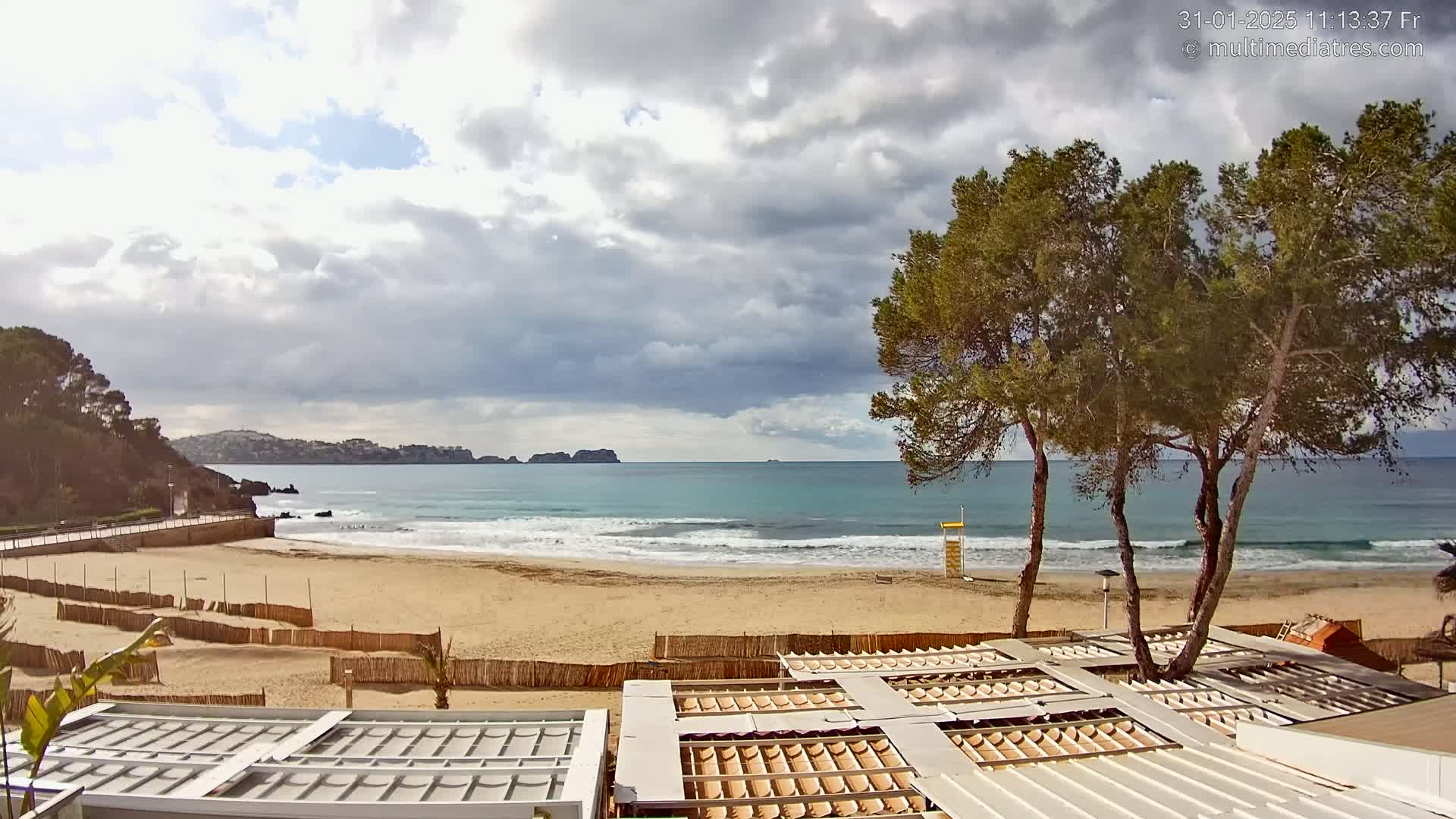 A sandy beach with gentle waves under a partly cloudy sky is partially enclosed by wooden fencing, with several covered structures and pine trees nearby.