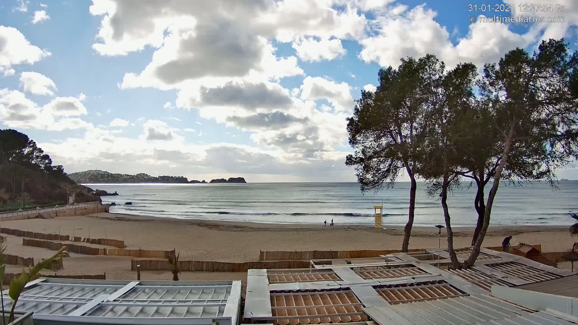 A sandy beach with gentle waves under a partly cloudy sky is viewed from above a beachfront structure.