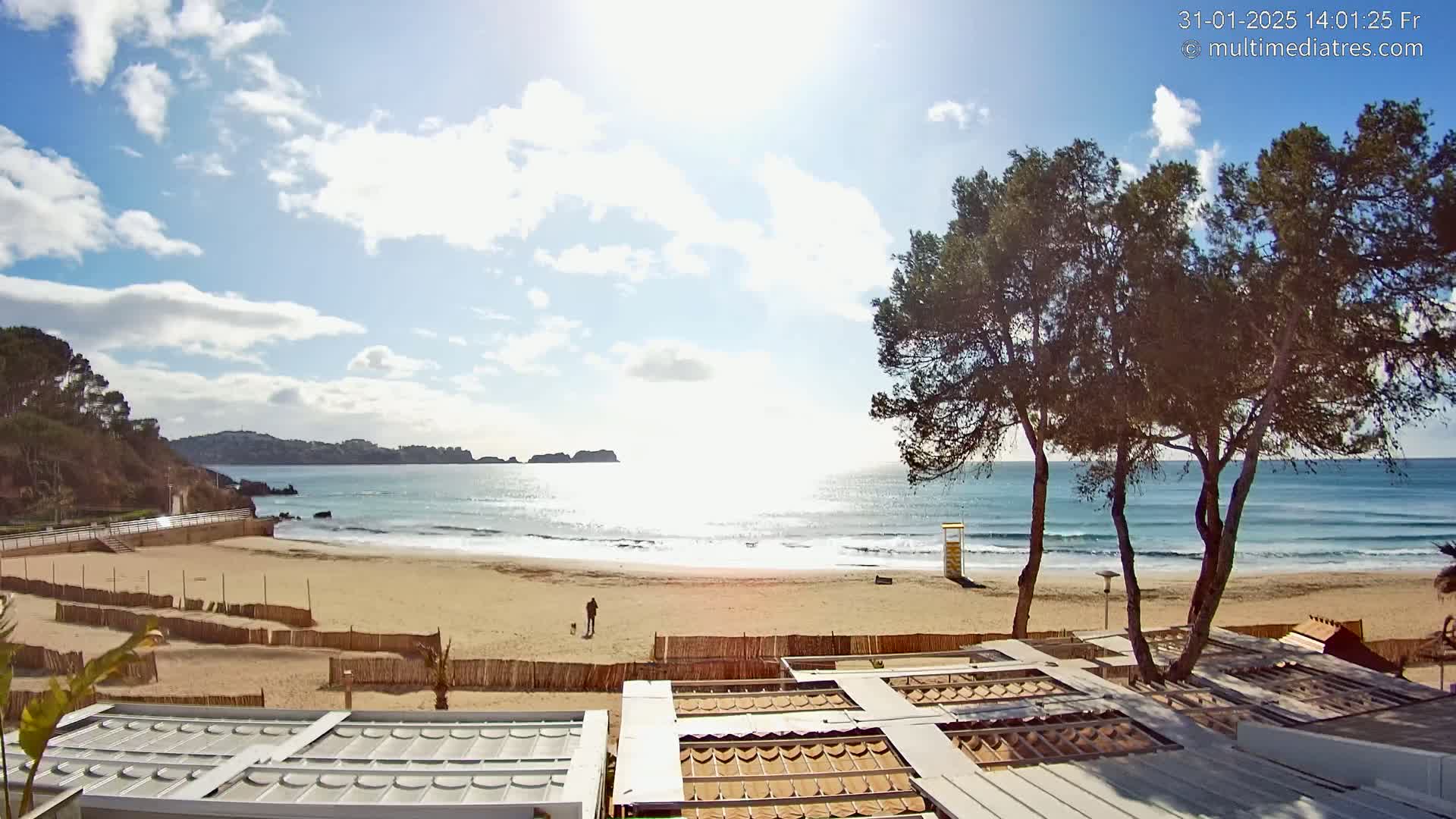 A sandy beach with gentle waves under a partly sunny sky, overlooked by several pine trees and beach structures.