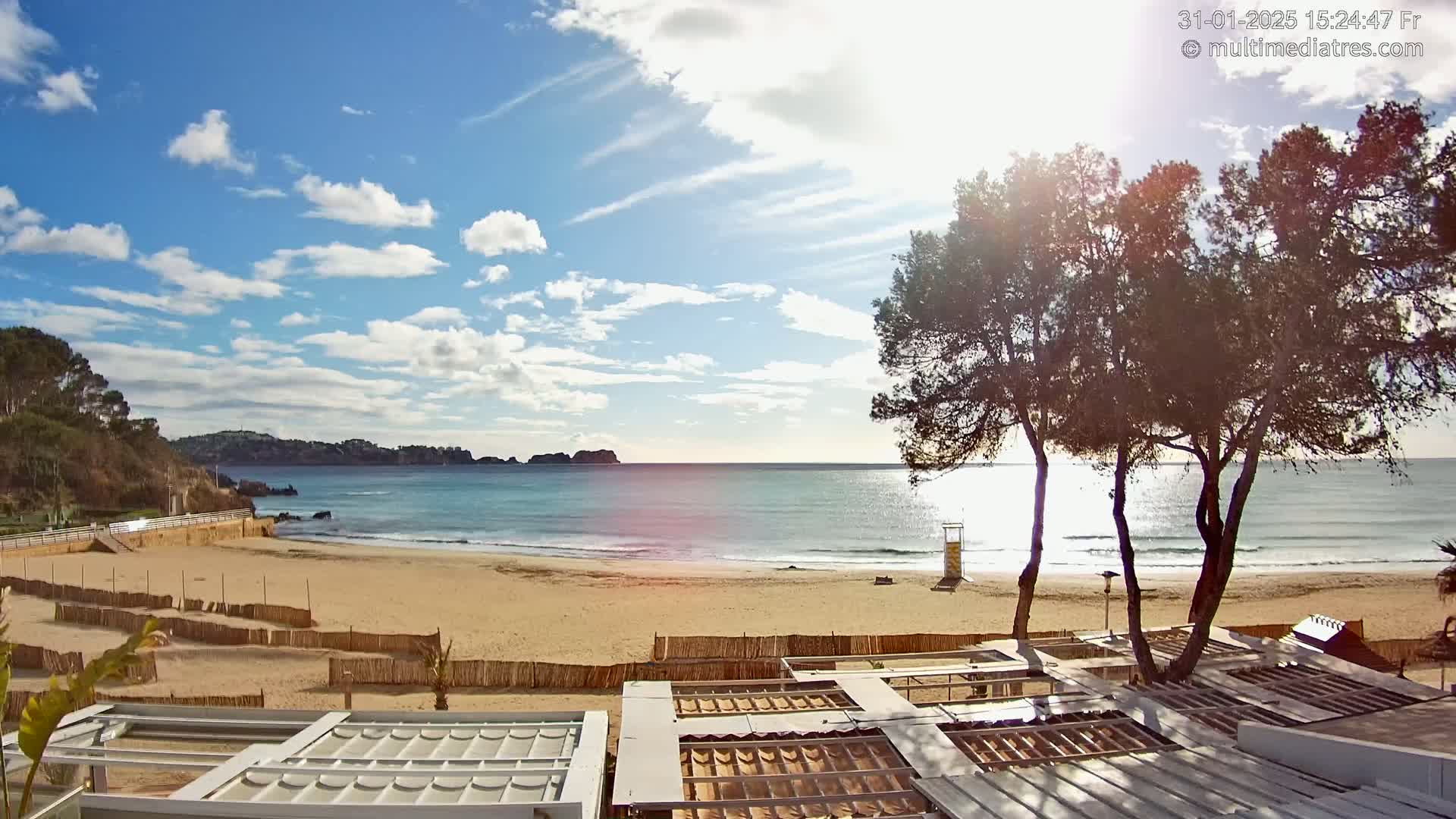 A sandy beach with calm, light-blue water under a mostly sunny sky with scattered clouds, viewed from behind a row of partially-constructed beachside structures.