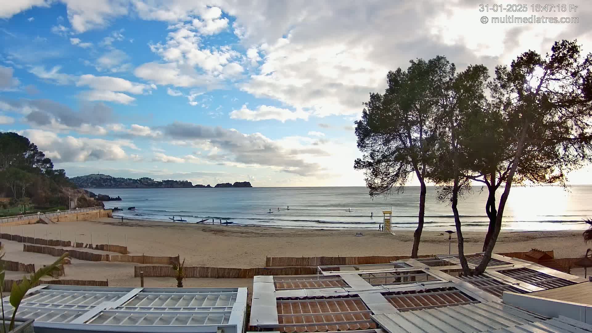A partly cloudy day at a beach with calm waves, several people surfing or paddle boarding, and trees in the foreground.