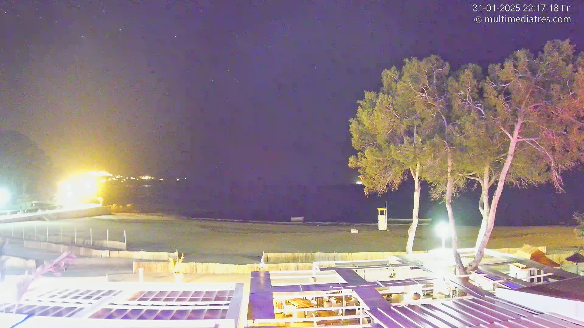 A nighttime view of a beach with a few trees and a building with a rooftop patio overlooking the calm water under a clear, starry sky.