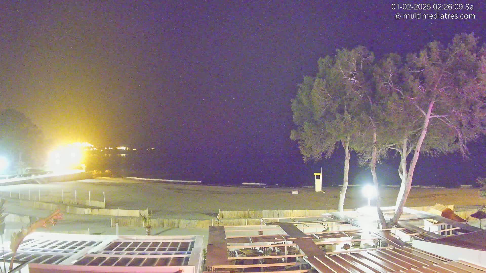 A nighttime view of a mostly empty beach, with a few trees and outdoor patio furniture visible in the foreground under a clear, dark sky.