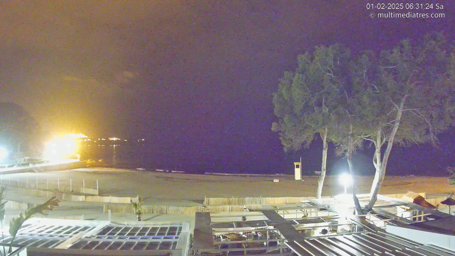 A nighttime view of a beach with calm waves, a lifeguard stand, and several trees, lit by artificial light, overlooking a building complex.