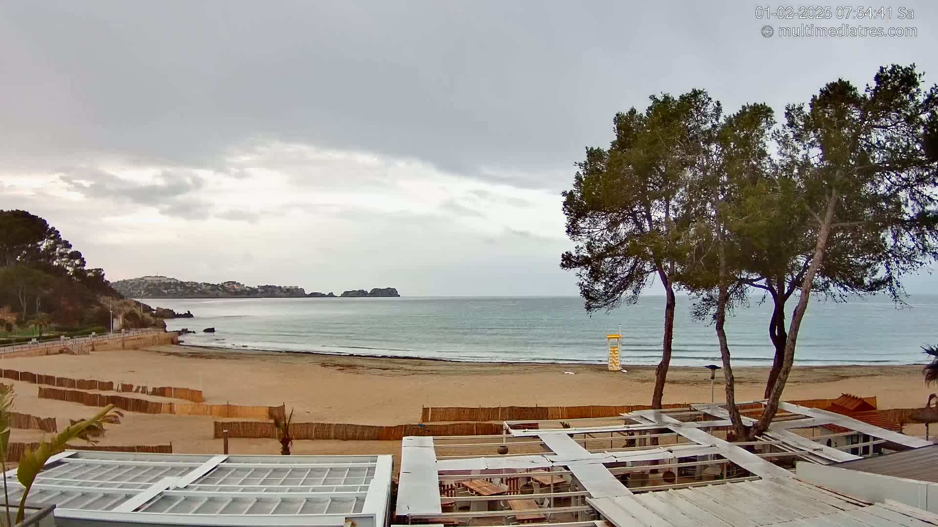 A sandy beach with calm ocean waves under an overcast sky is seen from above, with several pine trees and beachside structures in the foreground.