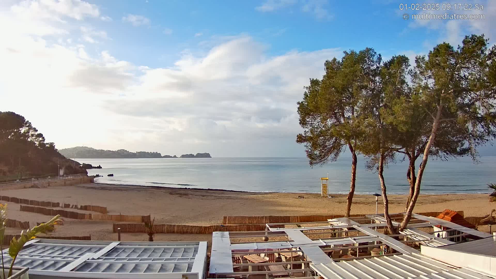 A sandy beach with calm ocean waters under a partly cloudy sky, viewed from above a beachfront patio area with several pine trees.