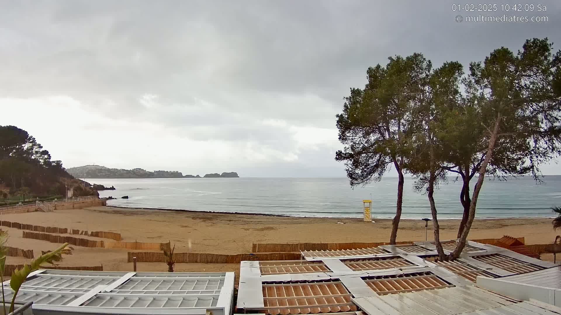 A sandy beach with calm waves under an overcast sky is bordered by vegetation, including several tall pine trees, and several beachside structures with open roofs.