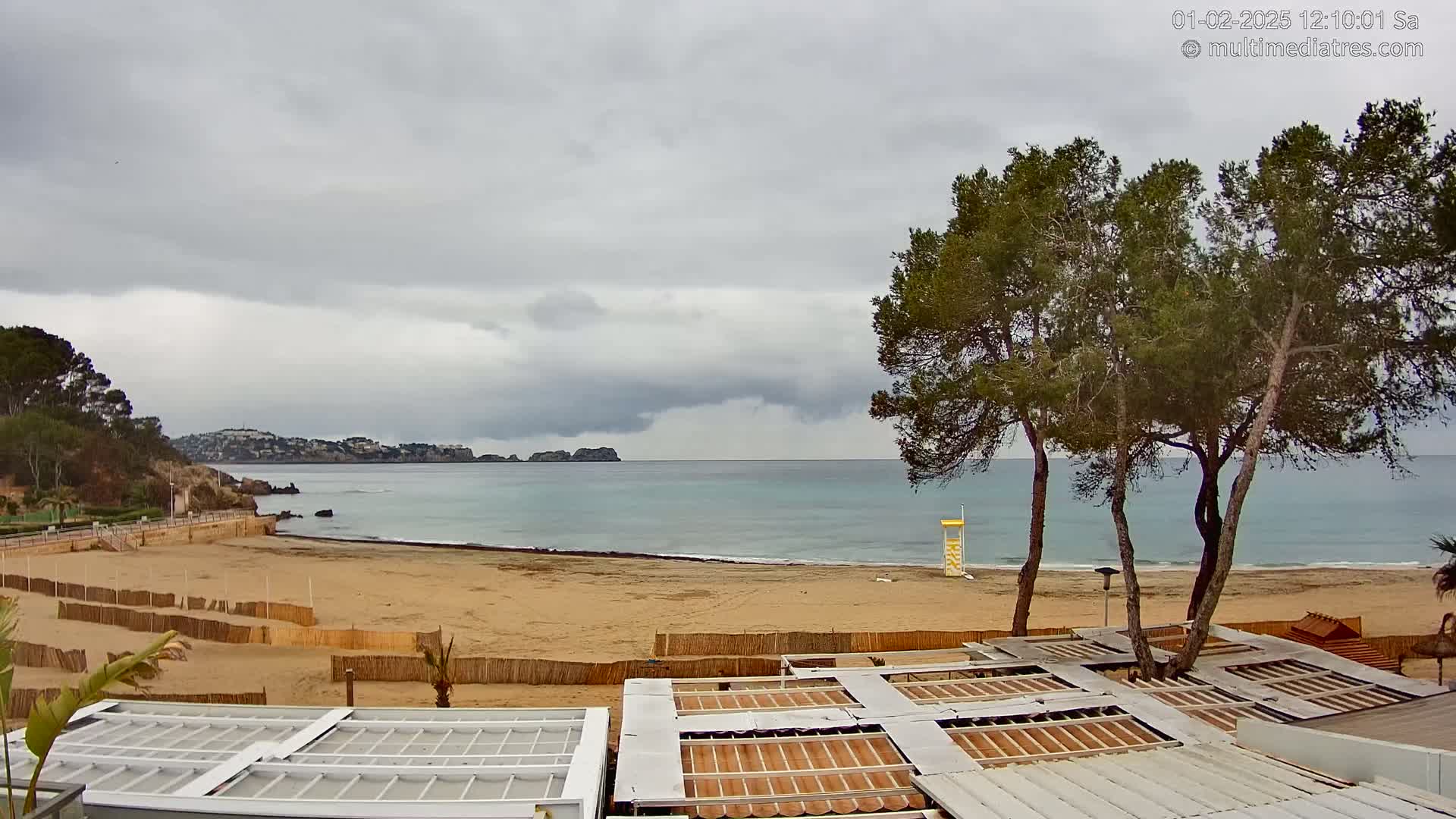 A sandy beach with calm water under an overcast sky is viewed from above a row of closed outdoor structures.