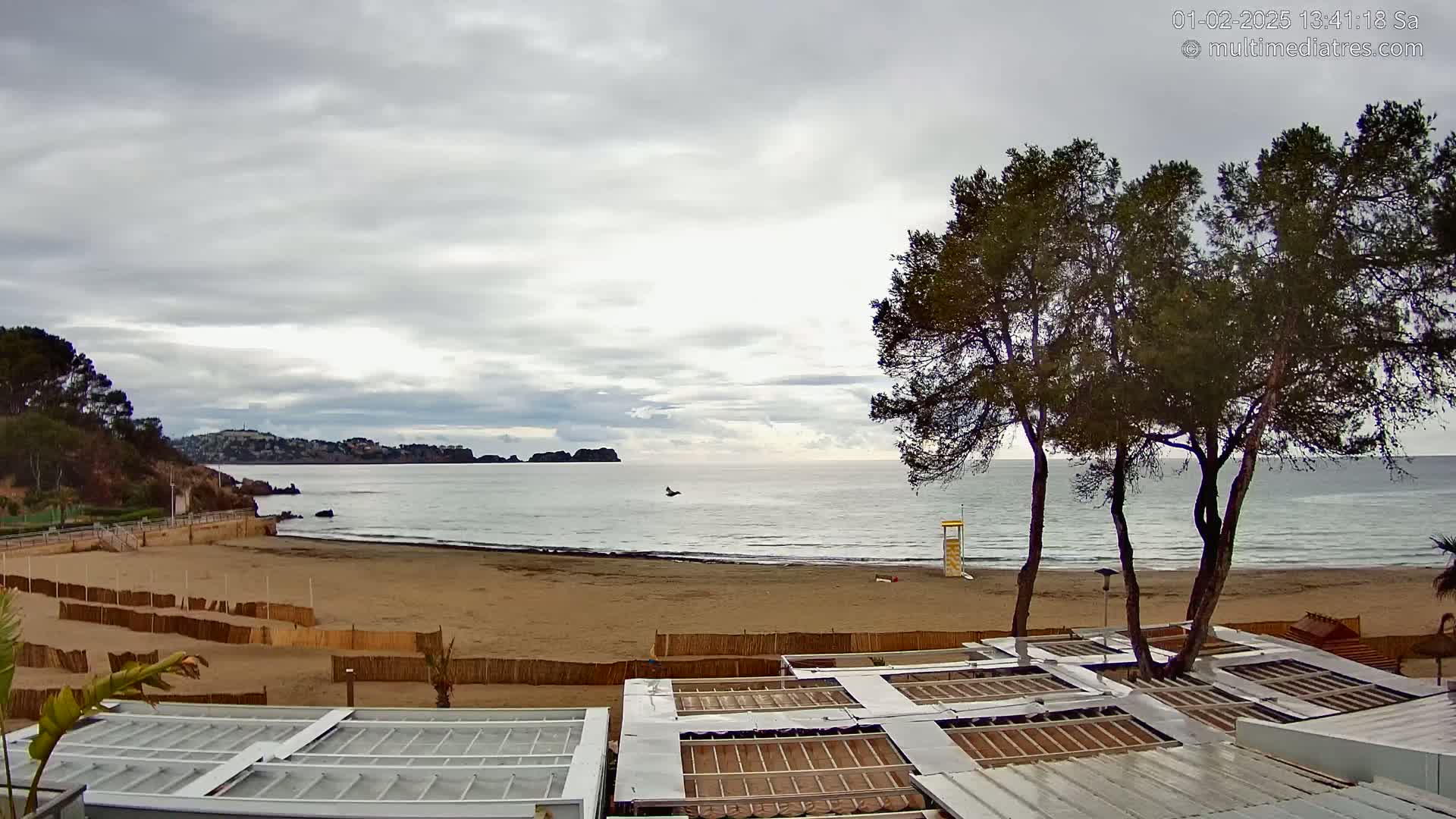 A sandy beach with calm ocean water under an overcast sky is viewed from above several structures with retractable roofs.