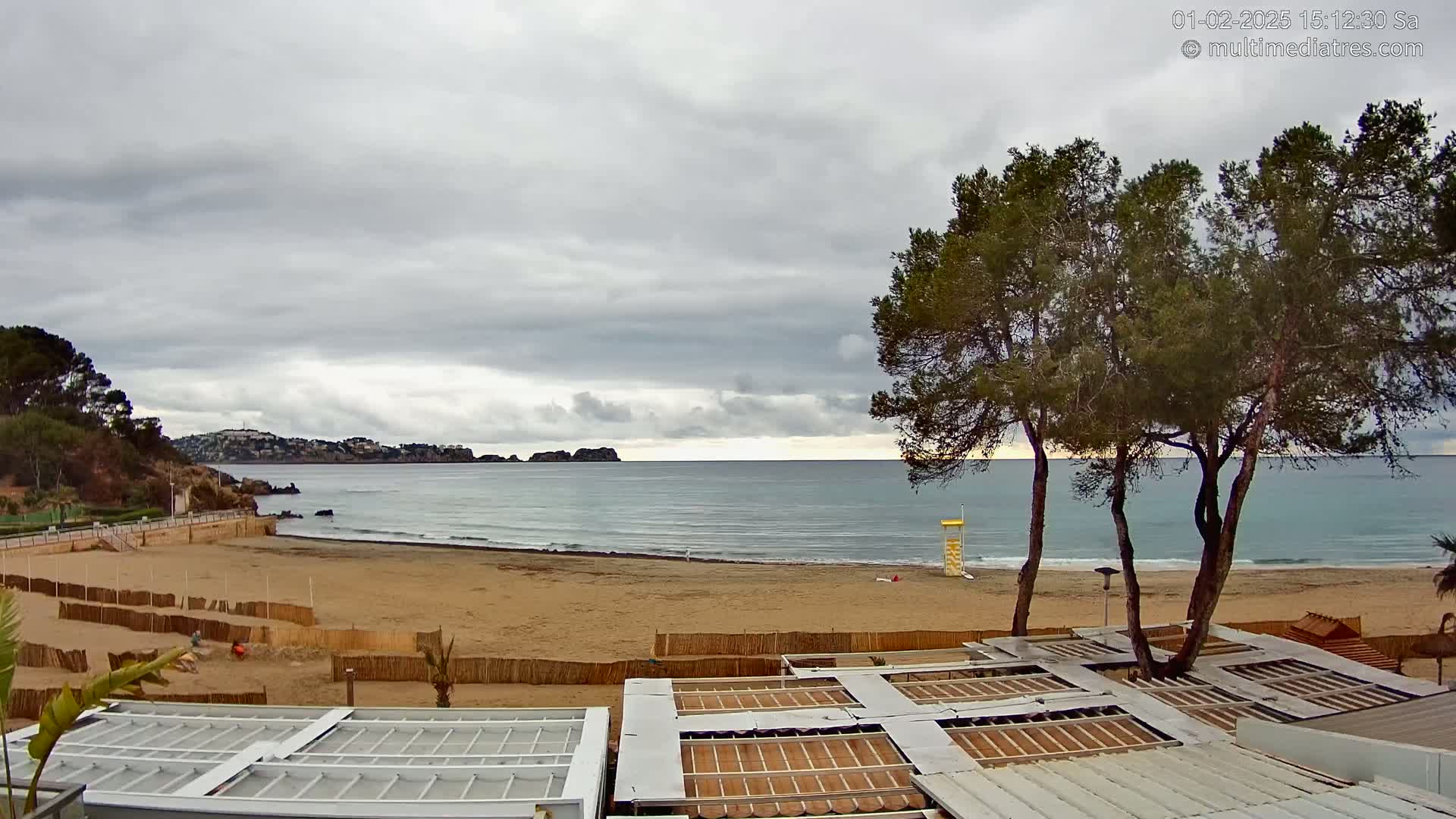 A sandy beach with calm waves stretches under an overcast sky, overlooked by several pine trees and partially covered beach structures.