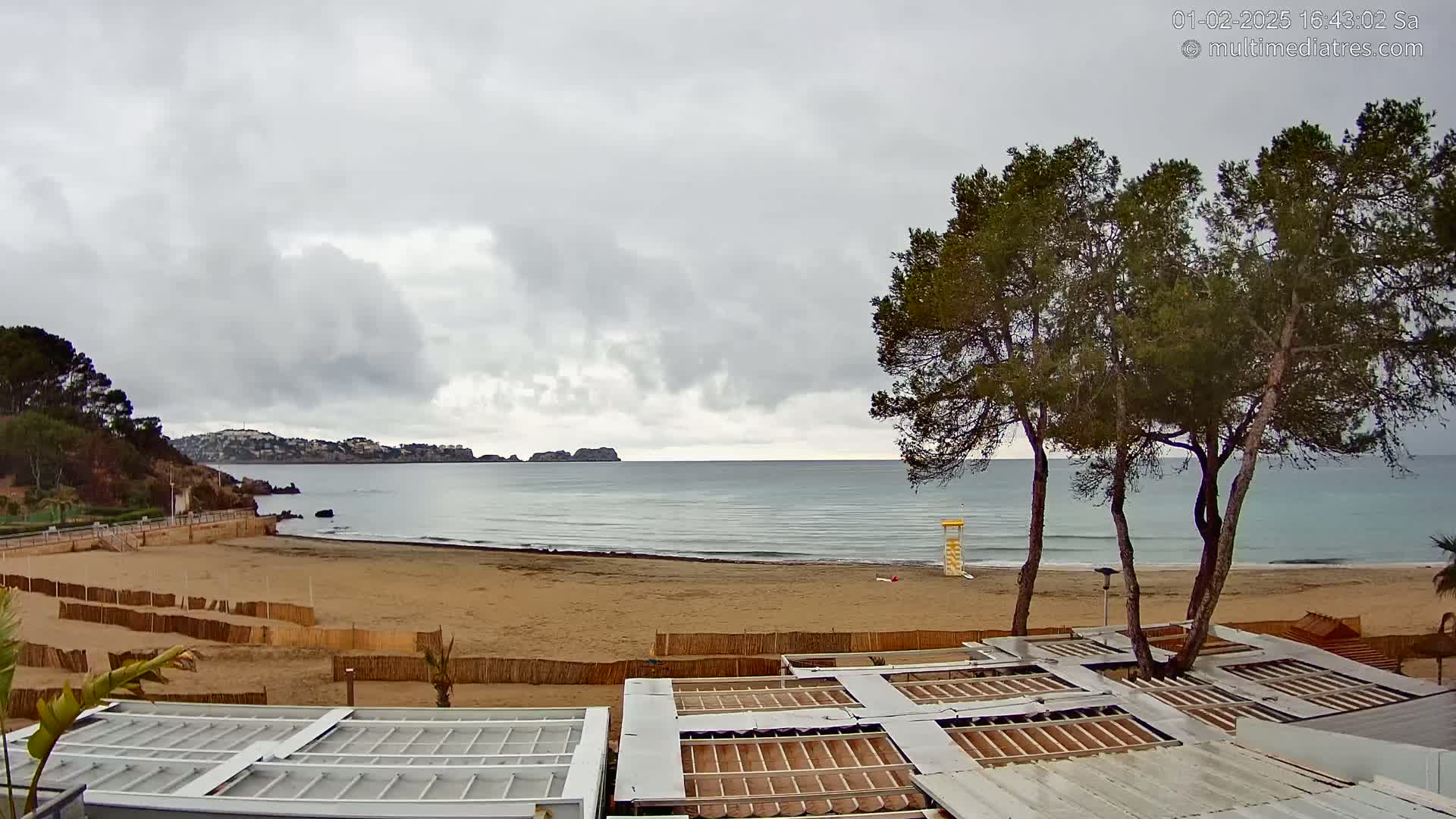 A sandy beach with calm ocean waves under an overcast sky is viewed from behind several beachside structures, with trees in the foreground and hills in the distance.