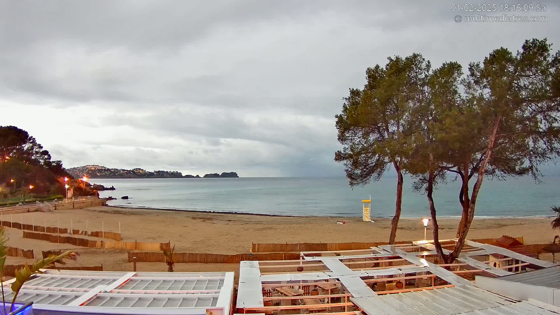 A sandy beach with calm ocean water under an overcast sky is viewed from above a beachside structure; several pine trees stand between the structure and the water.