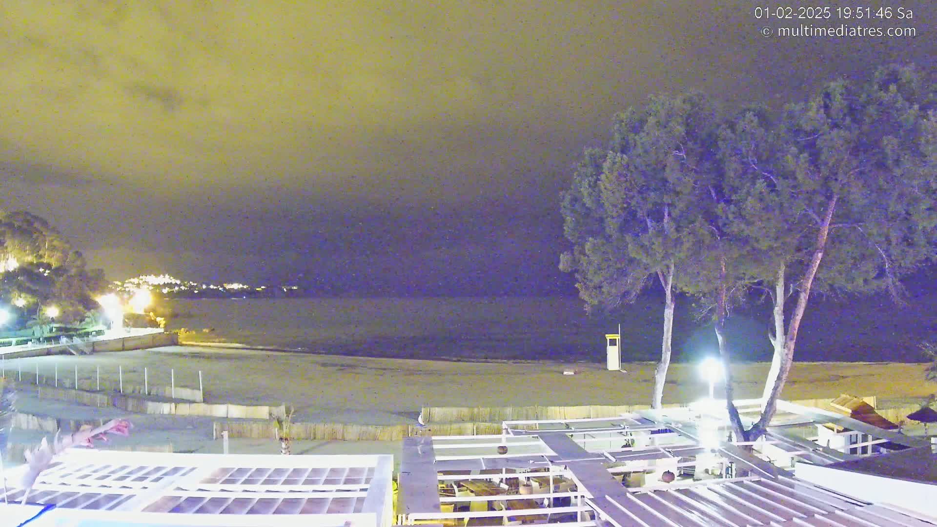 A nighttime view of a beach with calm water, some trees, and an outdoor structure with a partially visible roof.