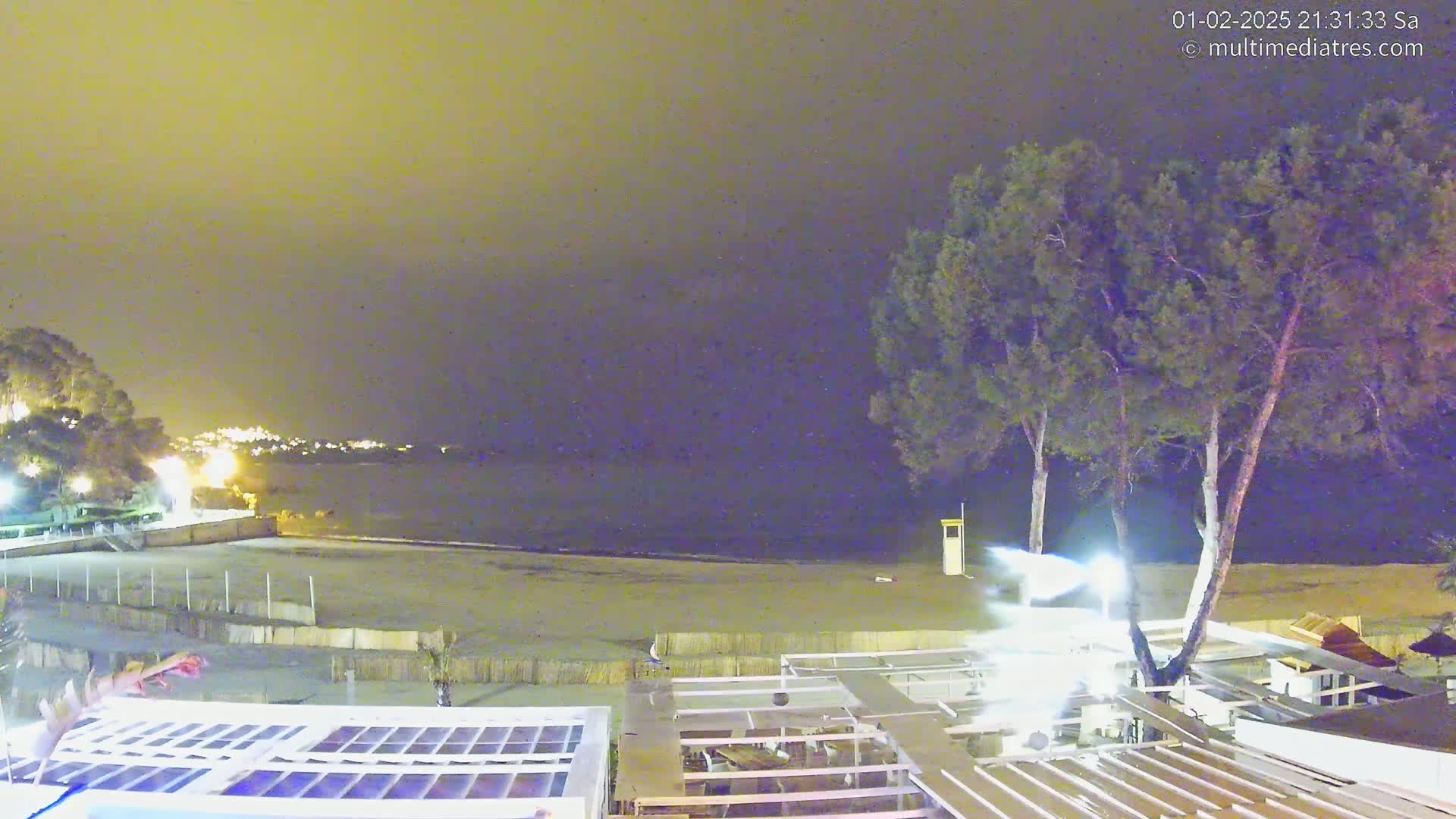 A nighttime view of a beach with calm water, a few lights visible in the distance, and several trees near a structure with a patio.