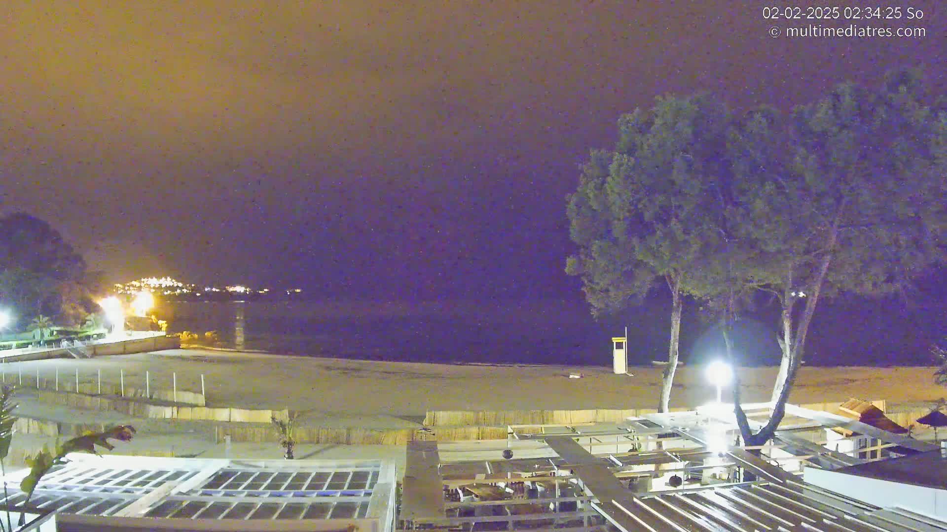 A nighttime view of a beach with calm water, illuminated by artificial lights, under a dark, possibly cloudy sky.
