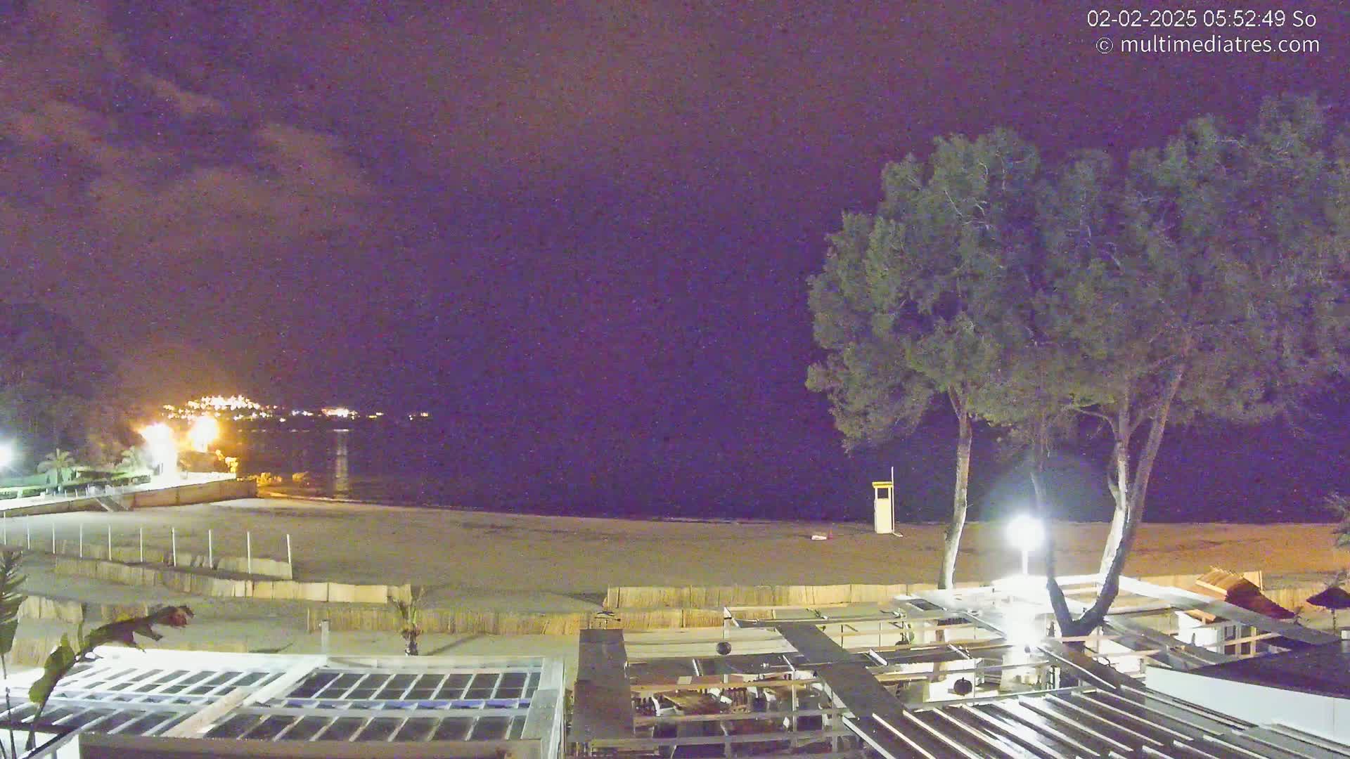 A nighttime view of a beach with calm water, two trees, and a partially visible beachfront establishment under a mostly clear, dark sky.