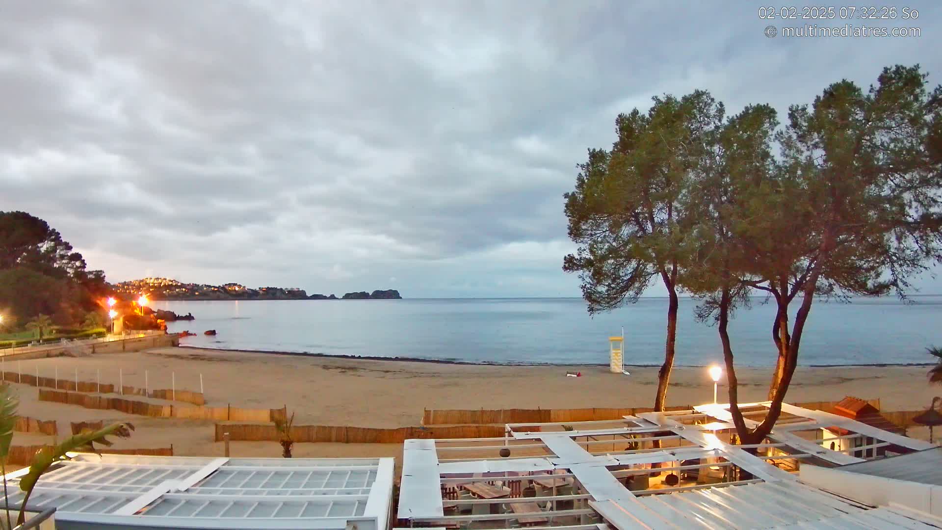 A sandy beach with calm water under an overcast sky is viewed from above a beachfront restaurant, with several tall pine trees in the foreground.