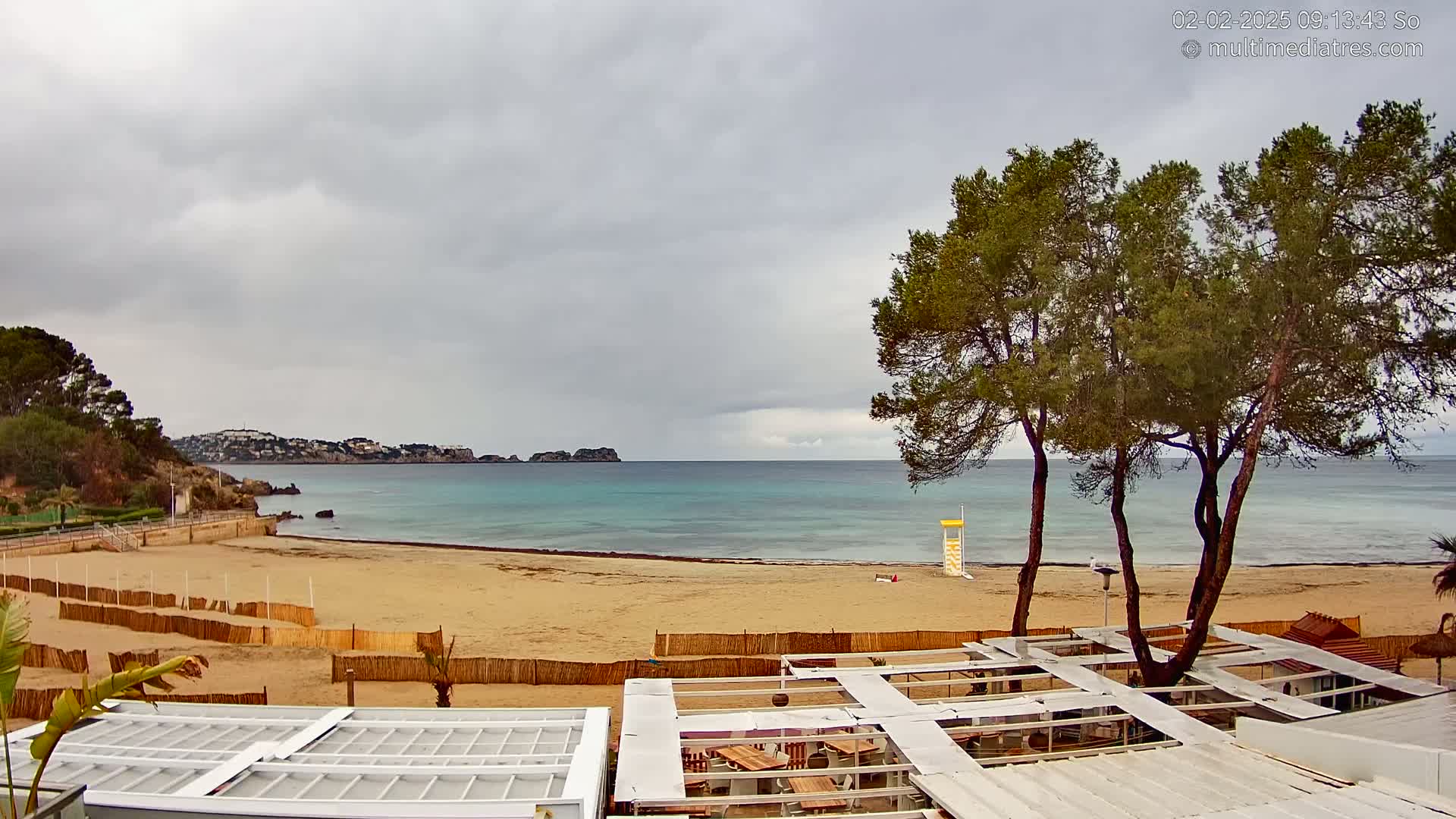 A sandy beach with calm turquoise water is partially enclosed by a wooden fence, with a lifeguard stand visible, under an overcast sky.