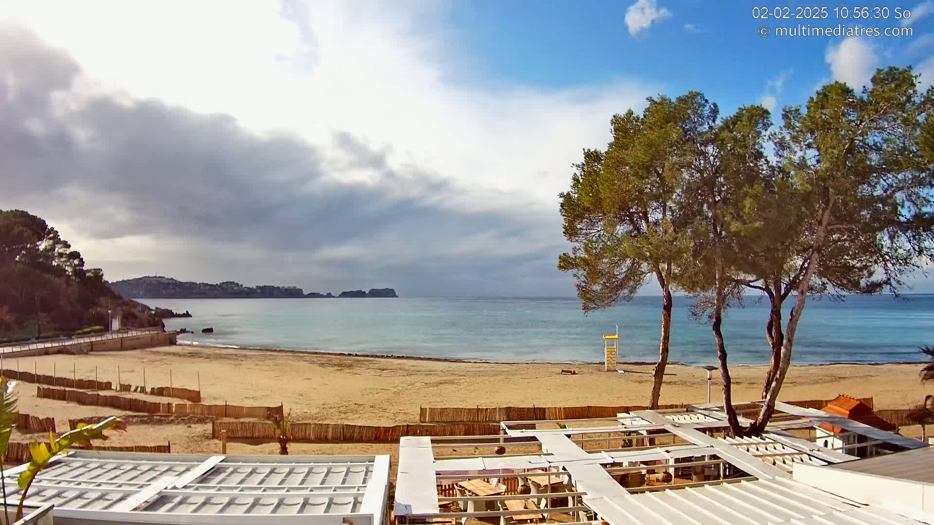 A partly cloudy day reveals a sandy beach with calm, blue water, several pine trees, and beachside structures.