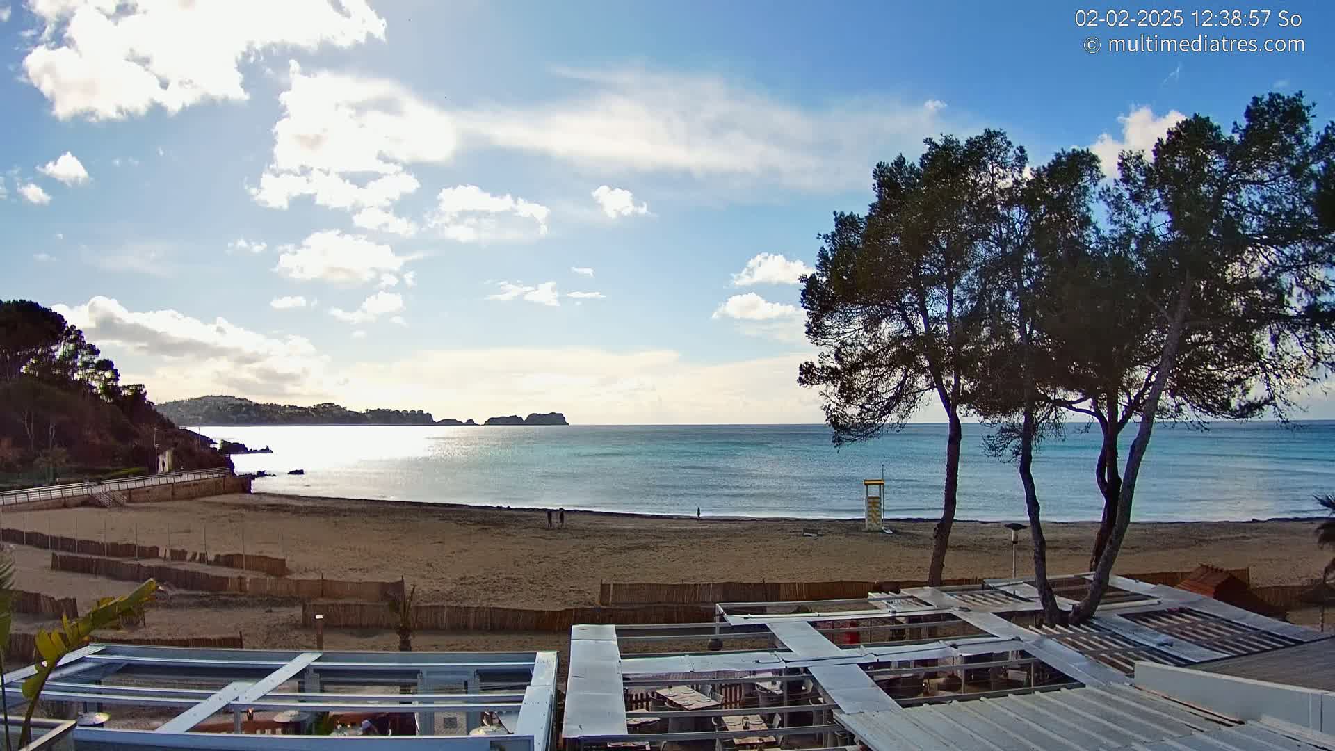 A sandy beach with calm, turquoise water stretches beneath a partly cloudy sky, framed by dark green trees and outdoor patio furniture.