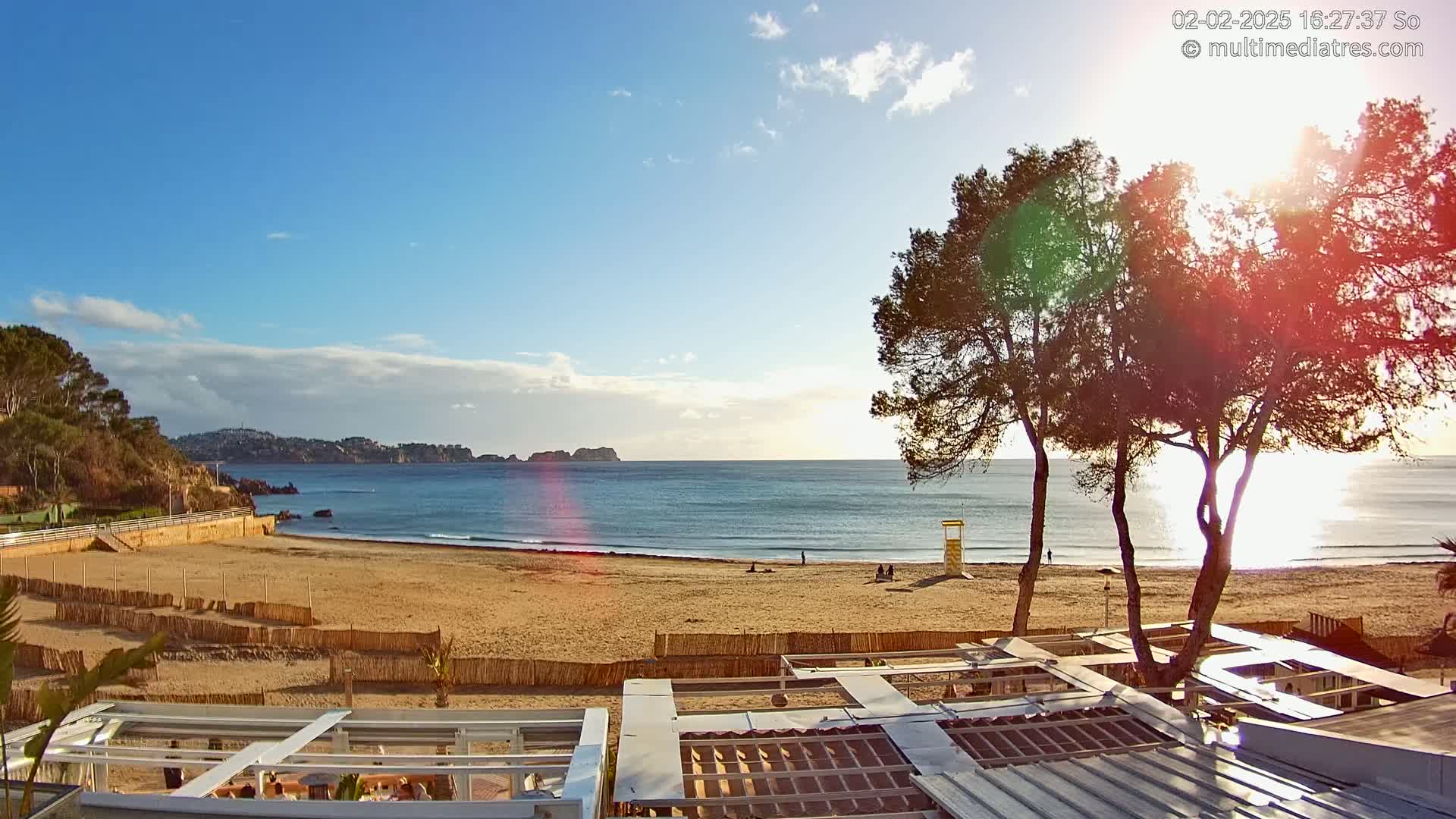 A sandy beach with calm ocean waves under a mostly sunny sky, partially obscured by trees in the foreground and hills in the distance.