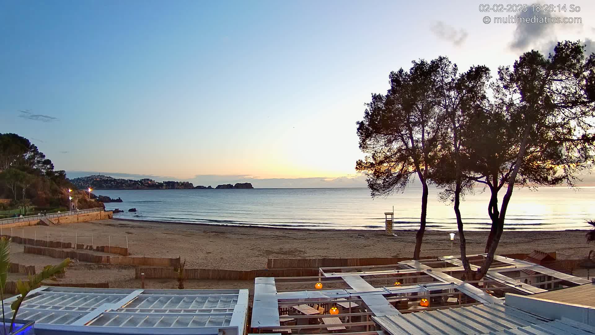 A sandy beach at sunrise, with calm ocean waves, is viewed from a beachside restaurant with a partially covered patio.