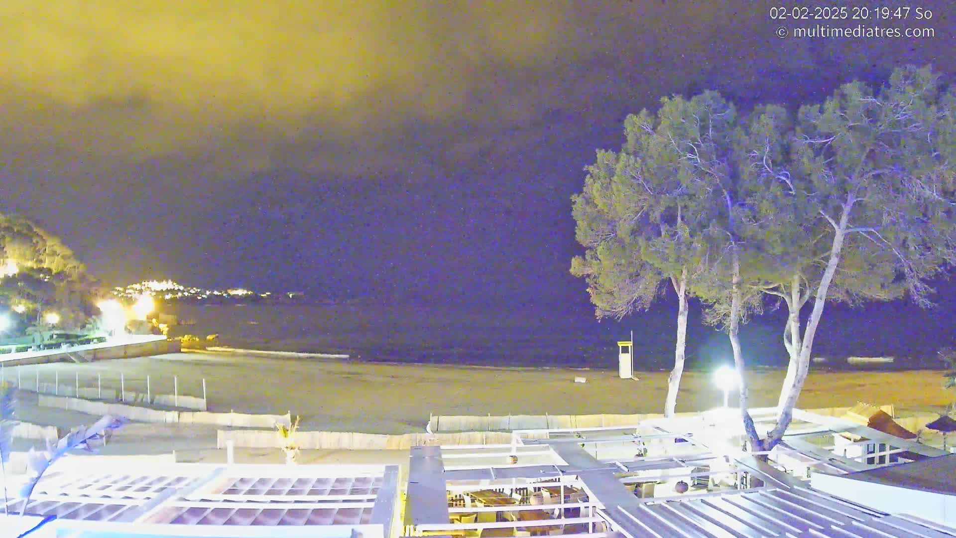 A nighttime view of a sandy beach, a calm sea, and a beachfront structure under a cloudy, yellow-tinged sky, with two tall trees standing near the structure.