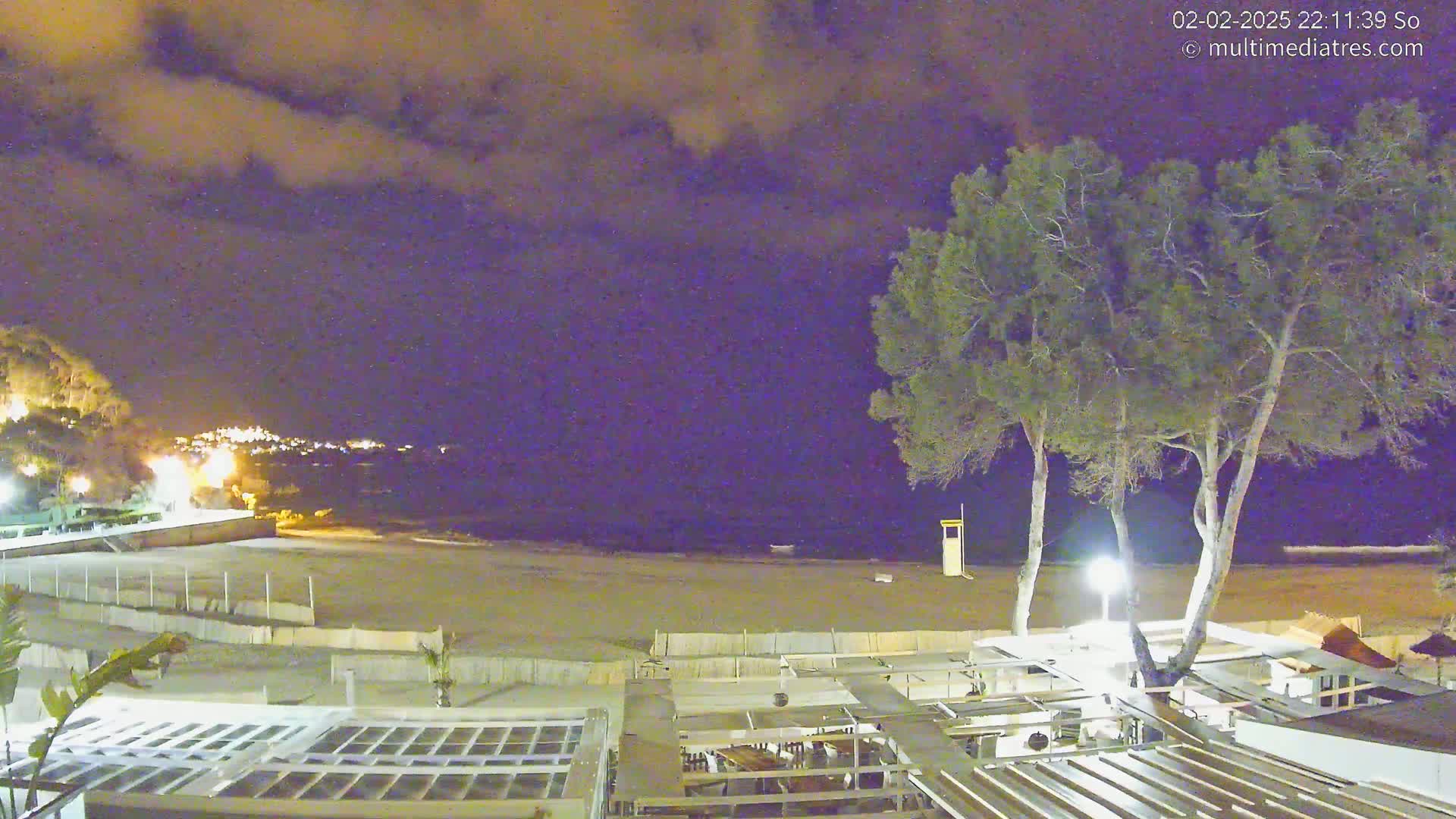 A nighttime view of a beach with a few trees, buildings, and lights under a cloudy sky.