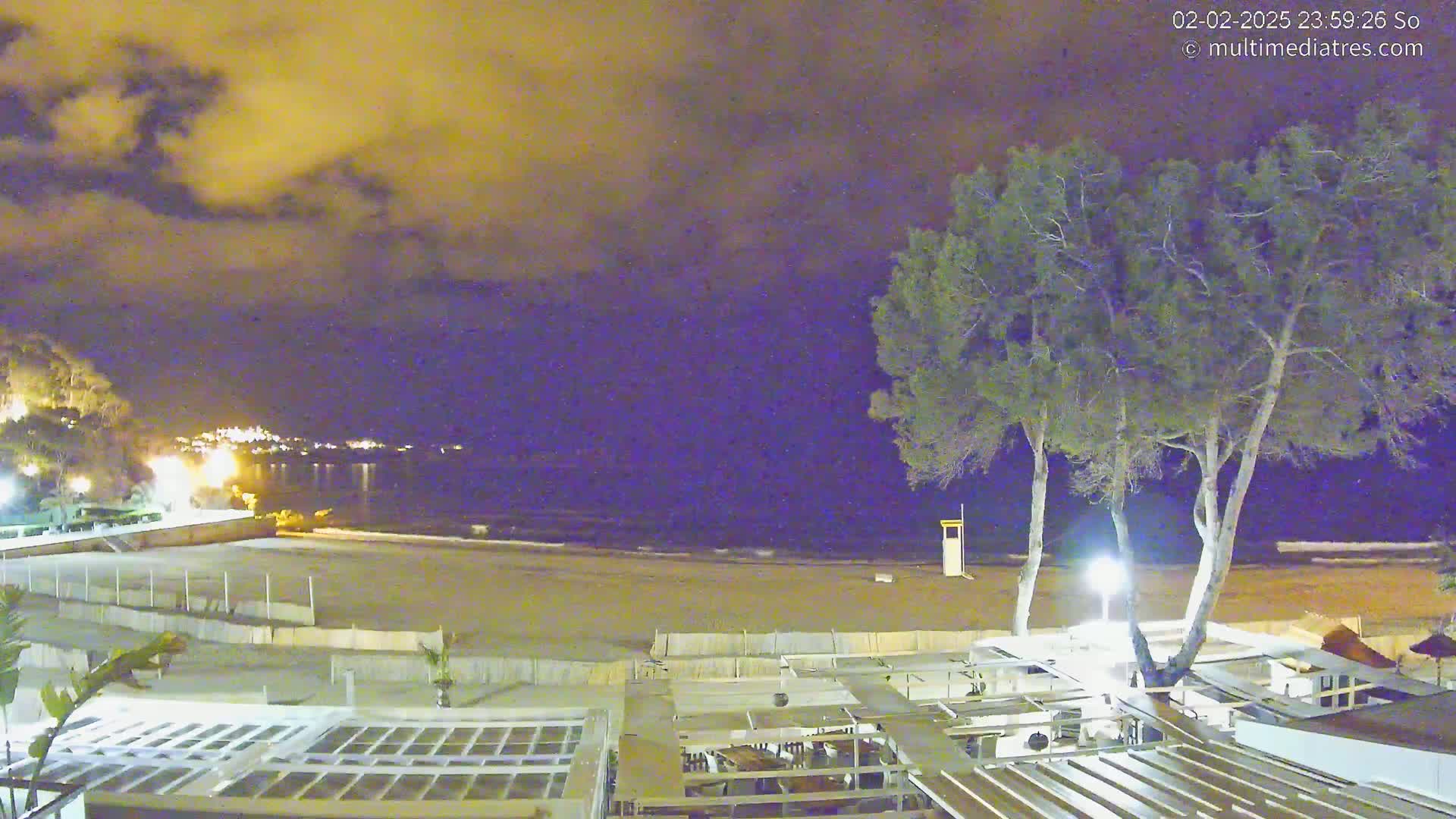 A nighttime view of a beach with a partially cloudy sky, showing a sandy shore, calm water, some trees, and outdoor seating areas near the water's edge.