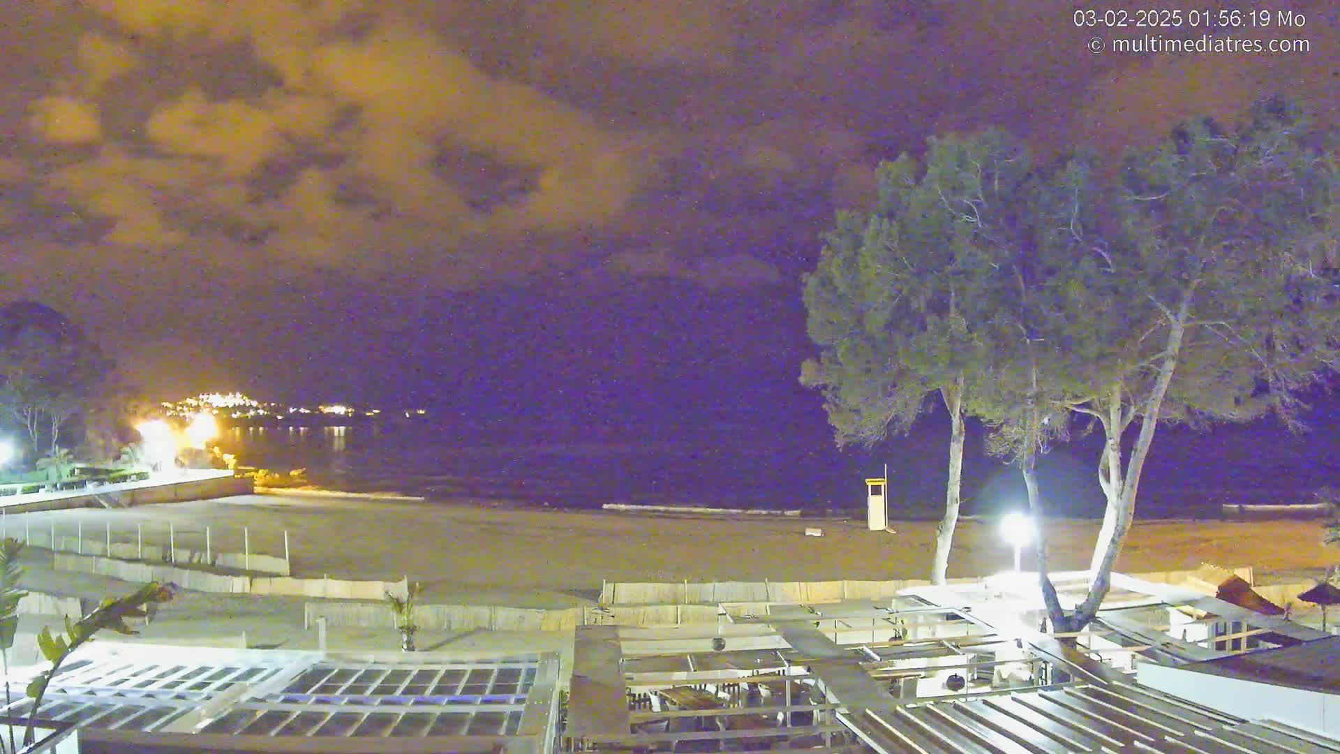 A nighttime view of a sandy beach, a calm ocean, and a shoreline with trees and outdoor structures under a cloudy sky.
