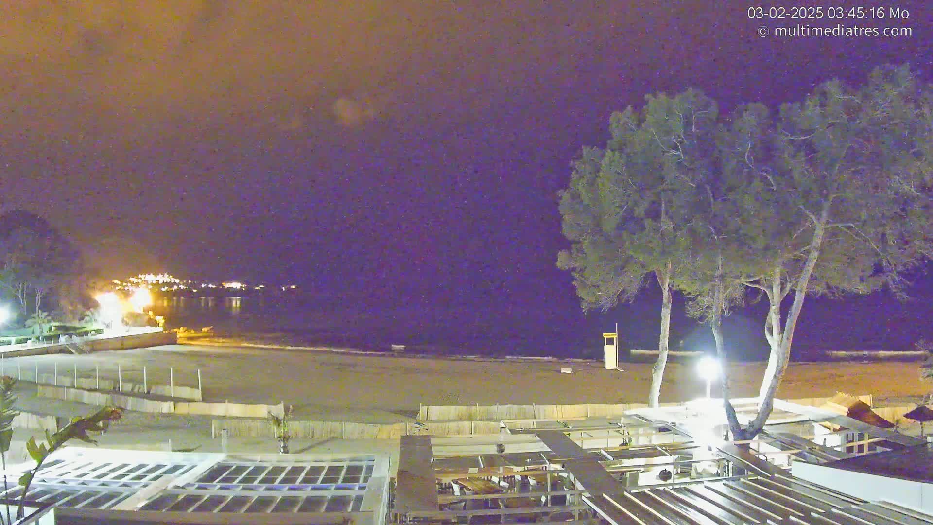 A nighttime view of a sandy beach with calm water, a few trees, and a partially visible beachfront establishment under a clear, dark sky.