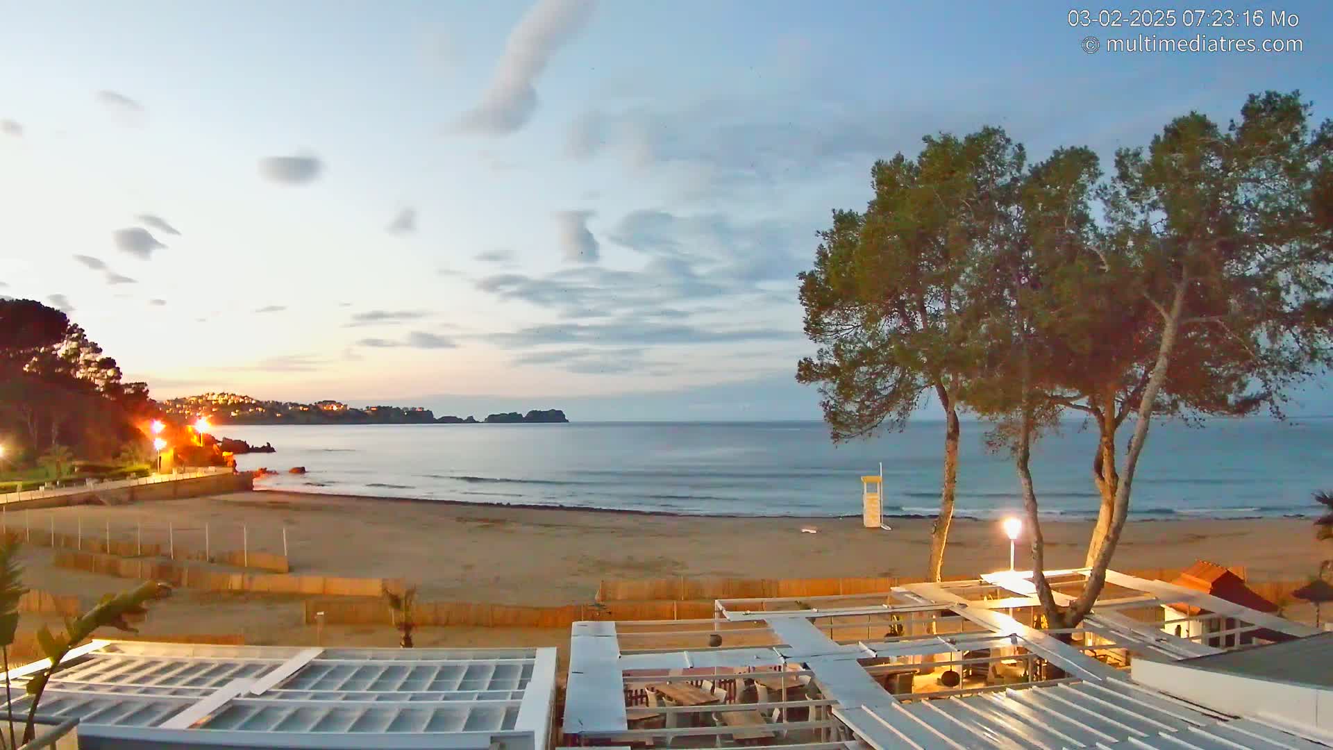 A sandy beach at dawn, under a mostly clear sky, is bordered by trees, buildings, and a calm ocean.