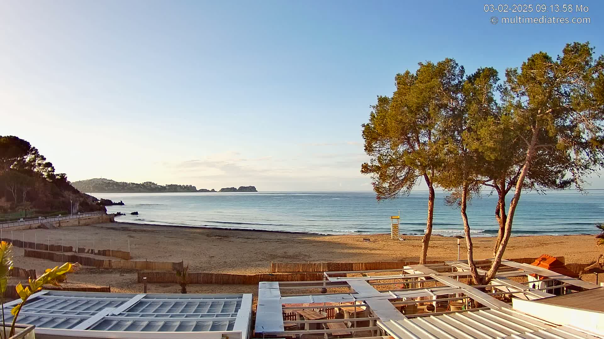 A sandy beach with calm ocean waves stretches towards a distant coastline under a clear, sunny sky, with several pine trees standing in the foreground near an outdoor patio structure.