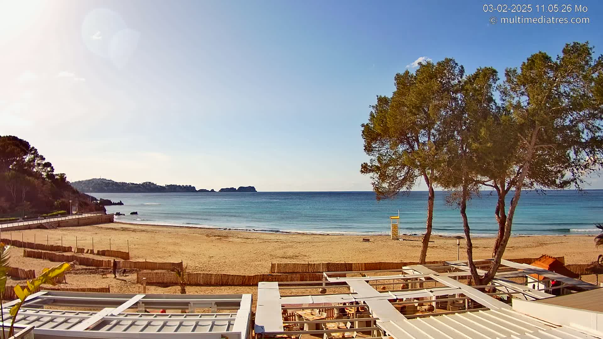 A sandy beach with calm, clear turquoise water is partially visible behind a beachfront restaurant with a white canopy structure under a sunny sky.