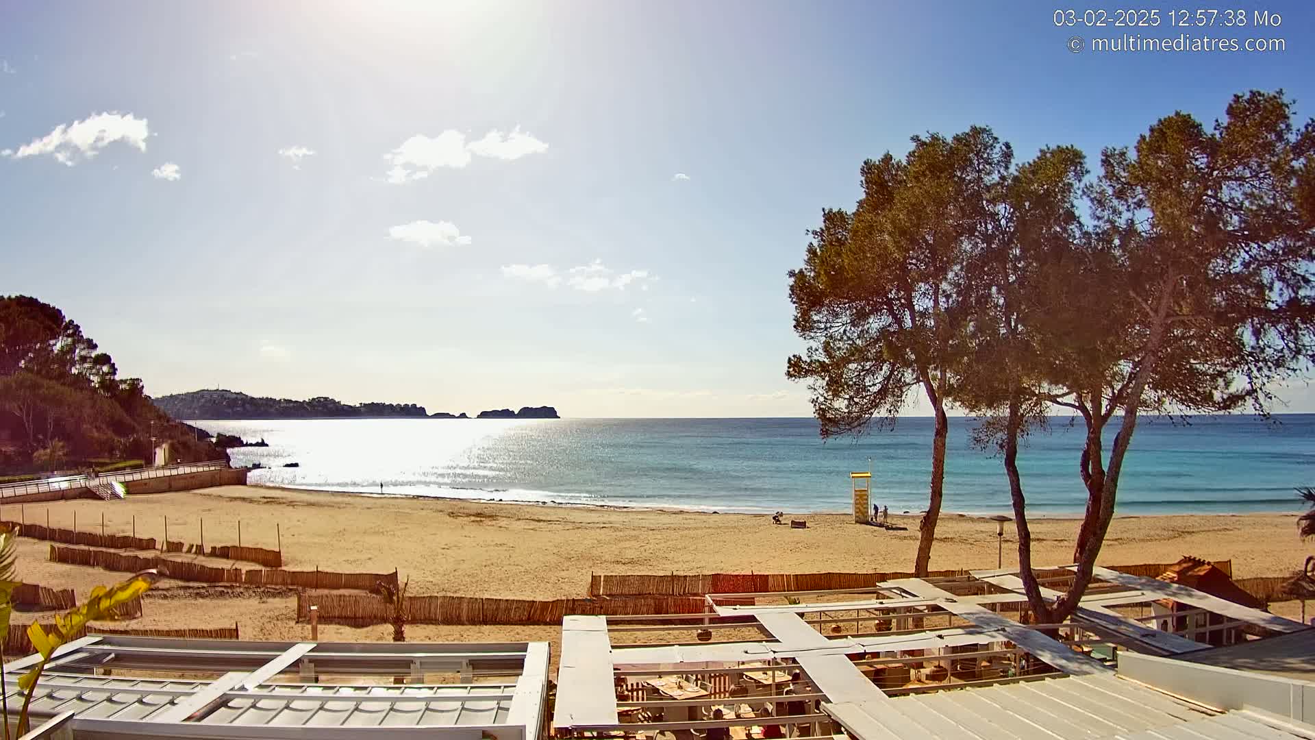 A sandy beach with calm, clear blue water stretches to a distant coastline under a mostly sunny sky, with a few clouds, and several pine trees framing the scene from an elevated viewpoint.