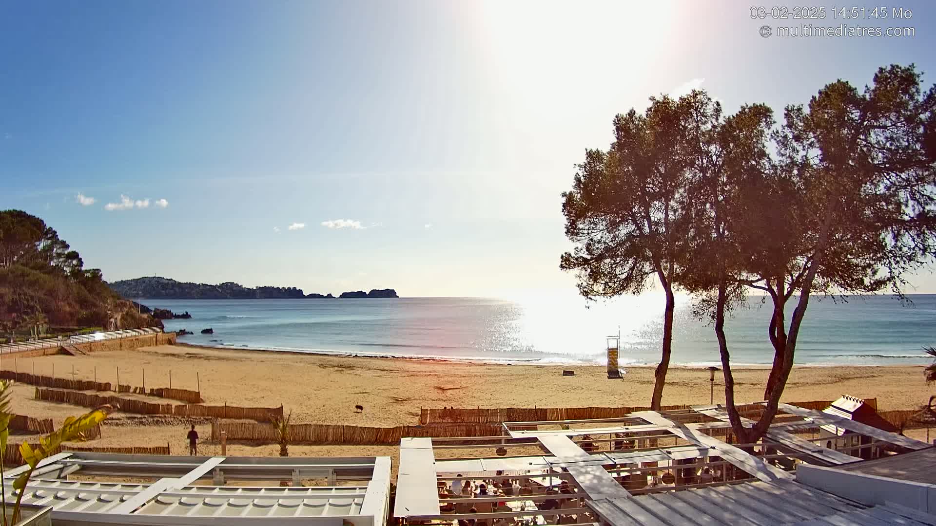 A sandy beach with calm ocean water, a few trees, and outdoor seating areas under a sunny sky.