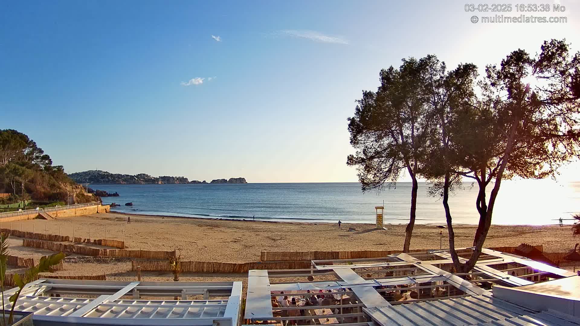 A sandy beach with calm ocean waves under a clear blue sky, viewed from behind a beachfront restaurant with several people dining.