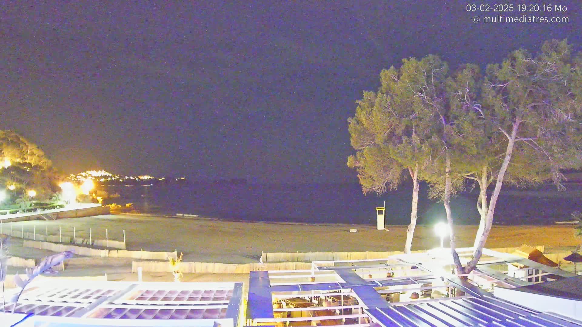 A nighttime view of a beach with calm water, a sandy shore,  trees, and a partially visible outdoor restaurant or bar area.