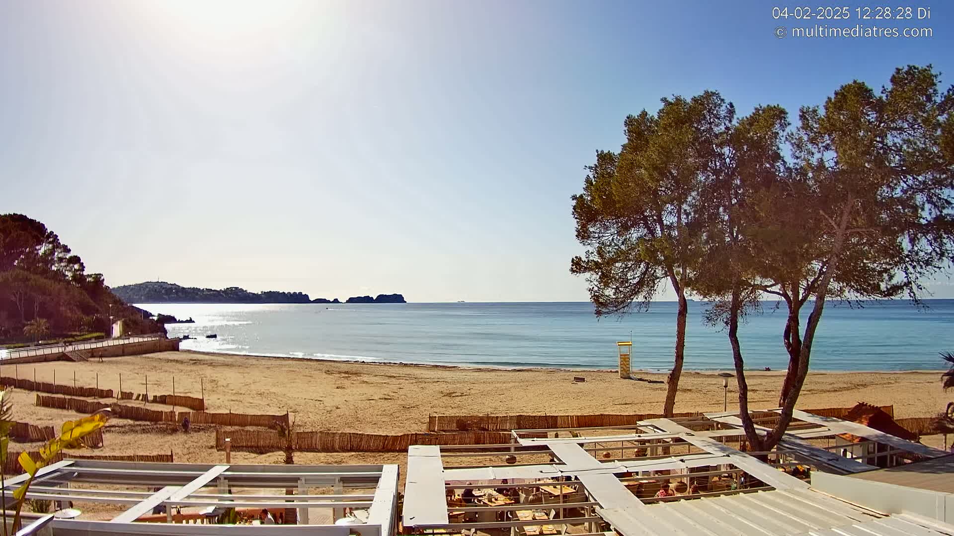 A sandy beach with calm, clear water stretches out under a sunny sky, bordered by a line of trees and a building with an outdoor seating area.