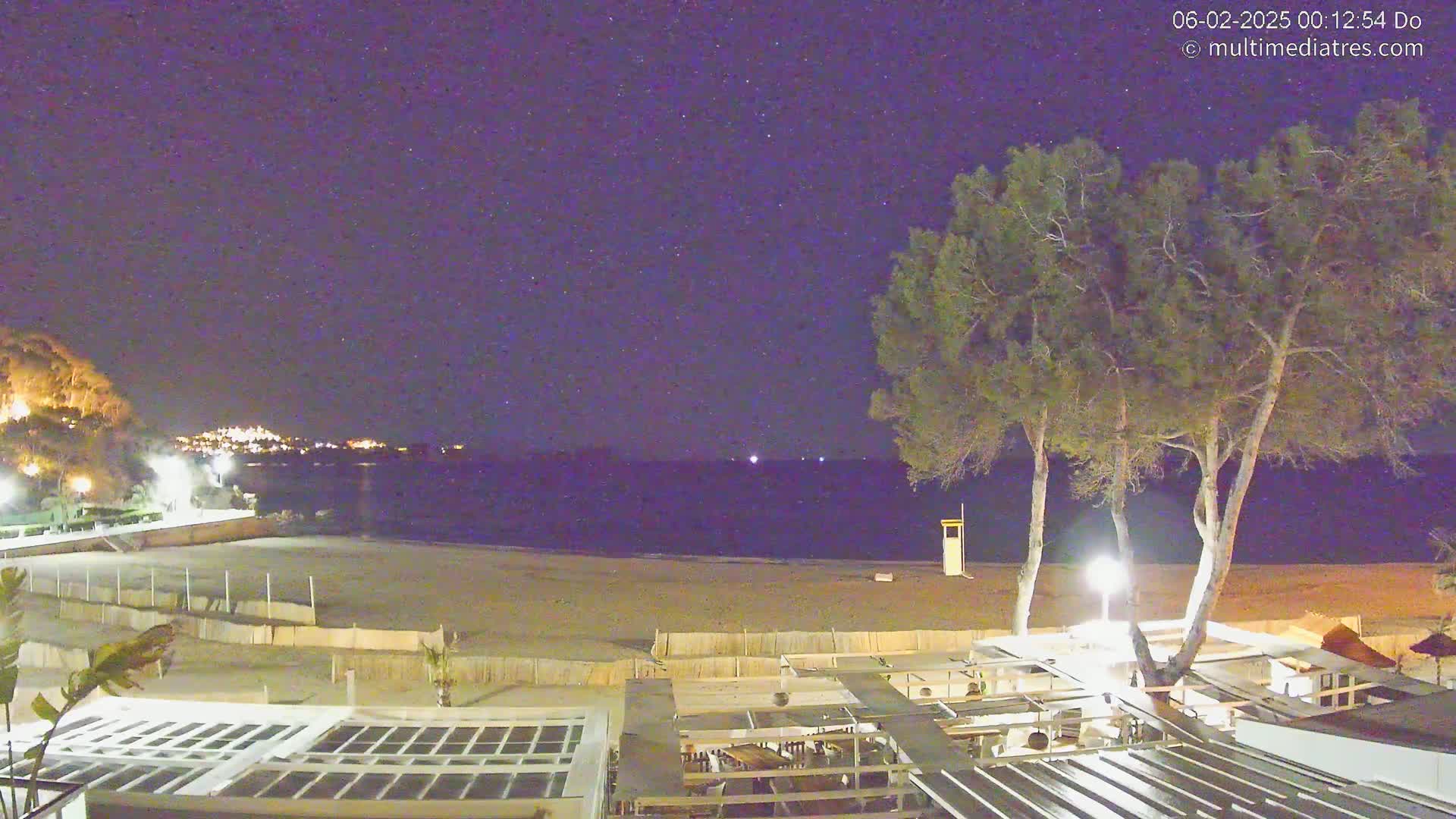 A nighttime view of a sandy beach, a calm sea, and a beachfront restaurant under a clear, starry sky.