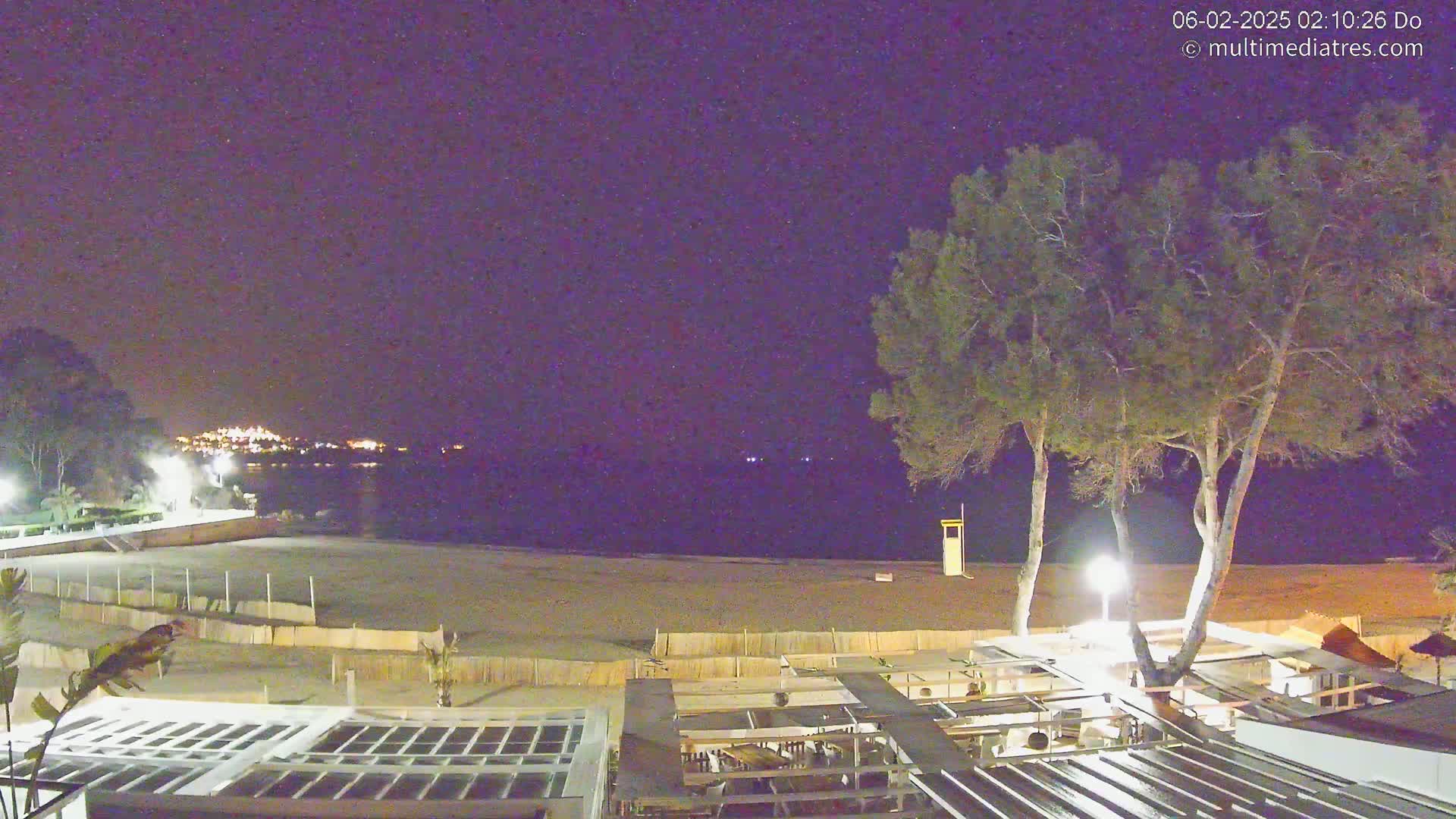 A nighttime view of a sandy beach with a building and trees, under a clear, dark sky.